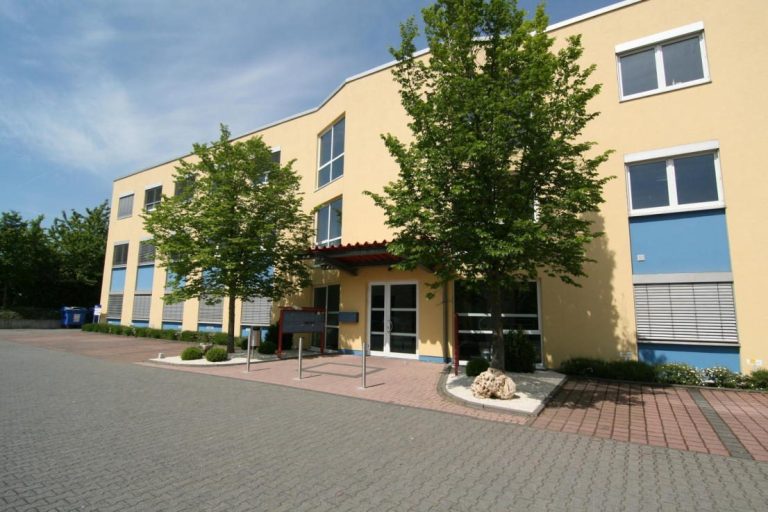 COMA PACPAGUNG GmbH Gelbes Gebäude mit großen Fenstern, umgeben von Bäumen und einem gepflasterten Gehweg.